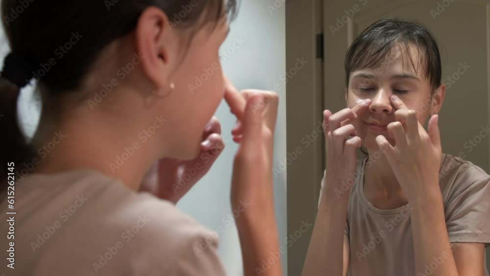 A young girl takes care of her skin. A cute teen girl applies cream on her face in front of a mirror in the bathroom. A young girl takes care of her skin. A cute teen girl applies cream on her face in front of a mirror in the bathroom.