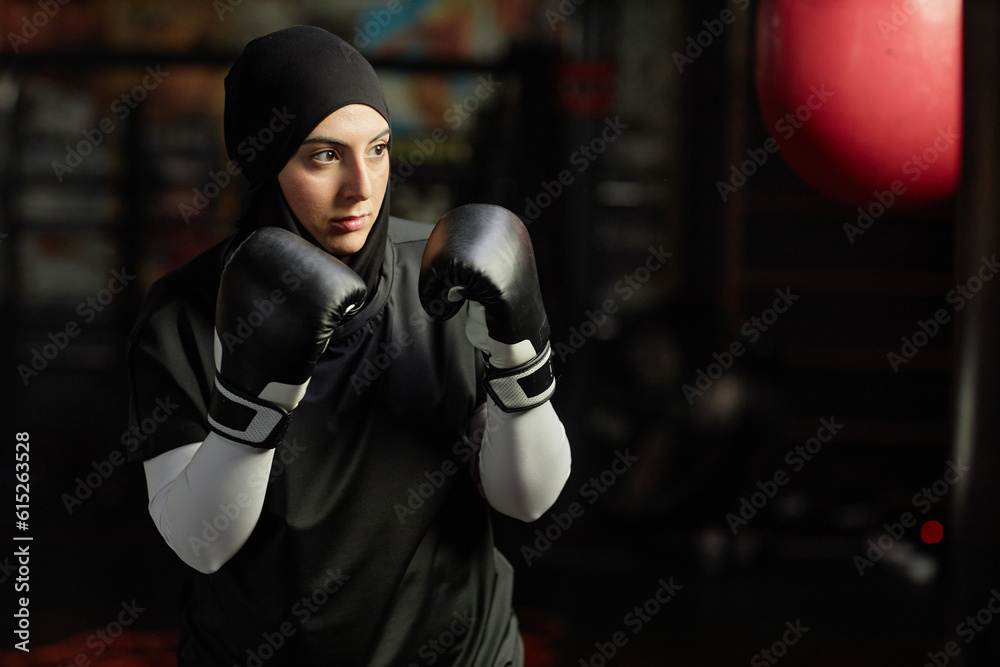 Strong young Muslim woman in boxing gloves, hijab and activewear looking at punching bag in gym and concentrating before hitting it