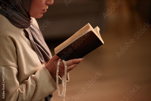 Close-up of young Islamic woman in beige pullover and grey headscarf holding open quran in front of herself while reading verses