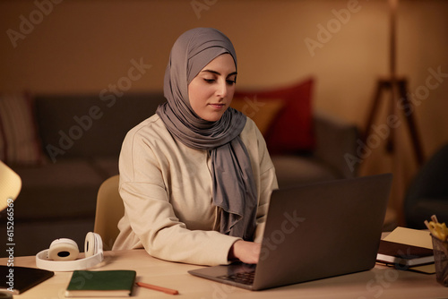 Young Muslim businesswoman in hijab and casualwear typing on laptop keyboard while sitting by workplace in home office and networking