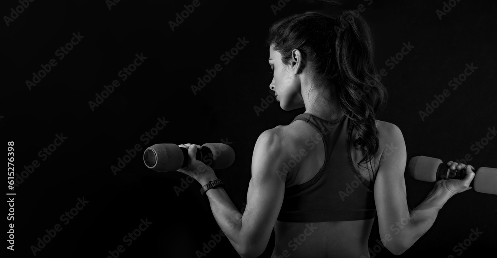 Female sporty muscular young serious woman doing strength workout on the shoulders, biceps and arms in sport bra holding dumbbells on black background. Closeup
