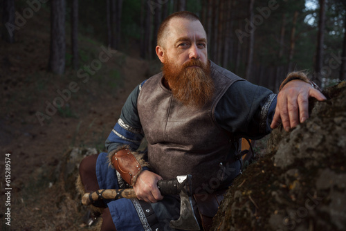 Portrait of medieval red-haired viking warrior with beard with an ax in forest