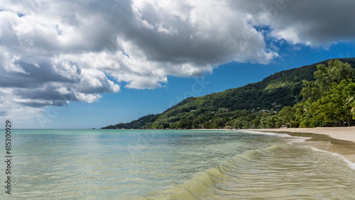 Fototapeta Naklejka Na Ścianę i Meble -  A wave of turquoise ocean rolls to the shore and spreads on the beach. Wet sand glistens. A hill overgrown with tropical vegetation, against a background of blue sky and clouds. Seychelles. Mahe.
