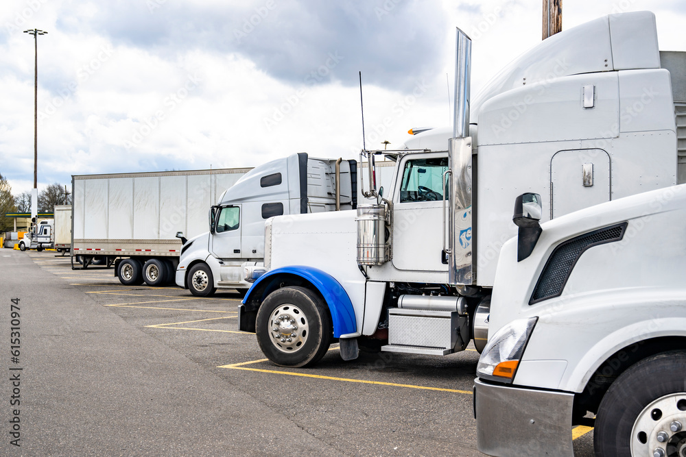 Different make and models of white big rig semi trucks with semi trailers standing in row on truck stop parking lot for truck driver rest Different make and models of white big rig semi trucks with semi trailers standing in row on truck stop parking lot for truck driver rest