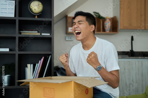 Young asian man unpacking  awaited parce looking inside, sitting at sofa, satisfied happy customer opening cardboard box with online store order, good shipping delivery service photo