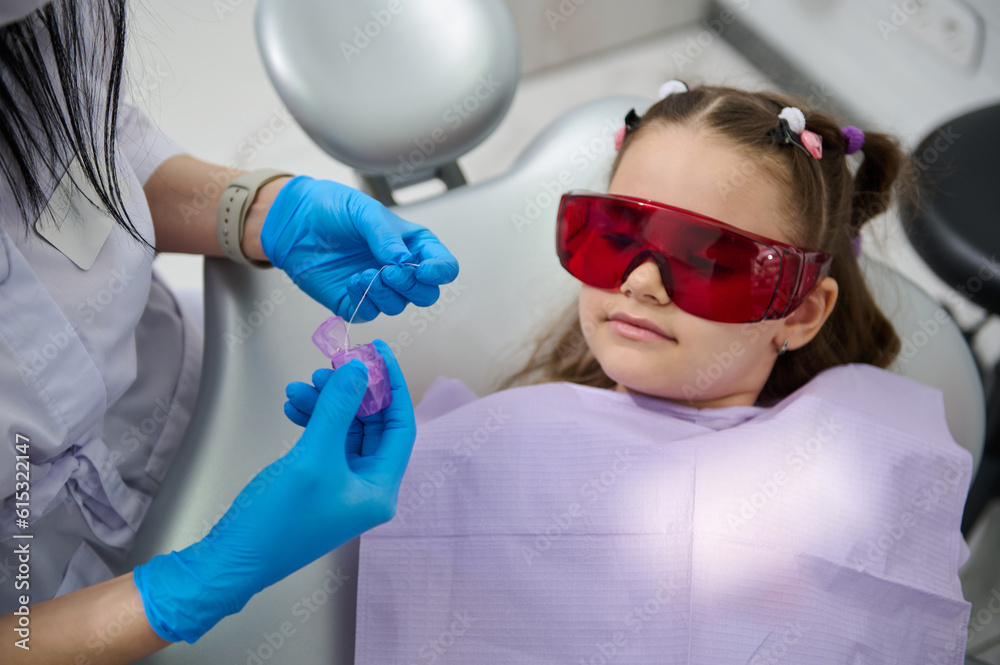 Naklejka premium Cute child girl listens to a dental hygienist explaining how to clean teeth using a dental floss after teeth brushing, during dentist's appointment in dentistry clinic. Oral care and hygiene concept