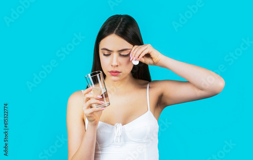 Girl take some pills, holds glass of water, isolated on blue. Young woman taking pill against headache. Brunette taking a pill with a glass of water. Woman taking drugs to releave headache photo