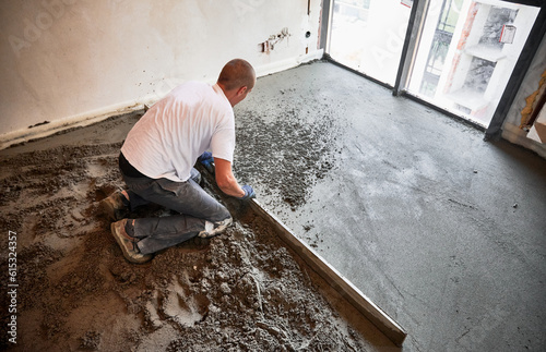Male construction worker using screed rail while screeding floor in living room. Man flattening and smoothing surface with straight edge in apartment during renovation. photo