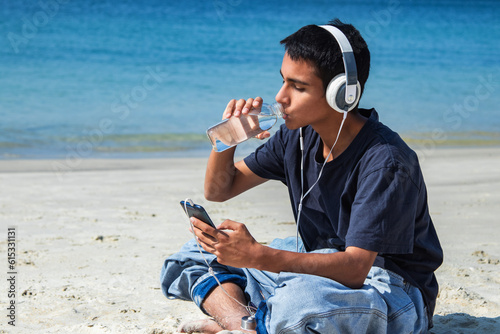 young man with headphones and mobile phone drinking water relaxed on the beach photo