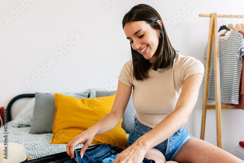 Young adult woman pack travel summer suitcase on bed for summer vacation or weekend getaway. Packing preparation before holiday departure photo