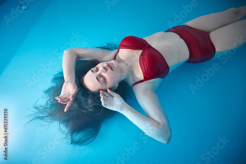 A young woman is swimming relaxed in a small pool, the blue background is visible through the clear water illuminated by light, dark hair, burgundy swimsuit, a day of relaxation at the spa.