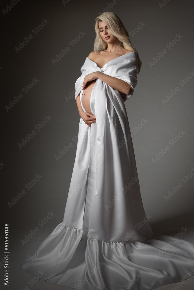 Pregnant Woman in White Dress looking at Belly. Beautiful Mother in Long Silk Gown over Dark Gray Background. Pregnancy Wedding Fashion