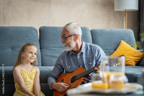 Grandfather and granddaughter sitting in the living room as he sings to her and plays guitar