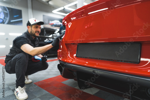 Professional male car detailer squatting next to the back of expensive red sports car and applying transparent vinyl foil to protect car paint. High quality photo