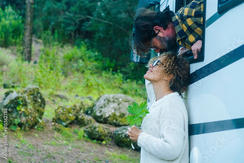 Happy adult cople have fun and love kissing outside a camper van parked in the green nature. Free parking in natural scenic place with trees. Alternative vacation and sweetness. Man and woman
