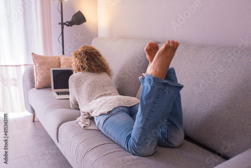 Back view of woman having total relax at home laying dowon on sofa and using laptop to surf the net and enjoy social media online activity. One female paople resting on couch in lazy sunday time photo