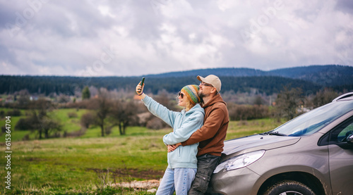 Romantic middle age couple hugging standing next to cars during auto travel in countryside. Taking a selfie using a smartphone