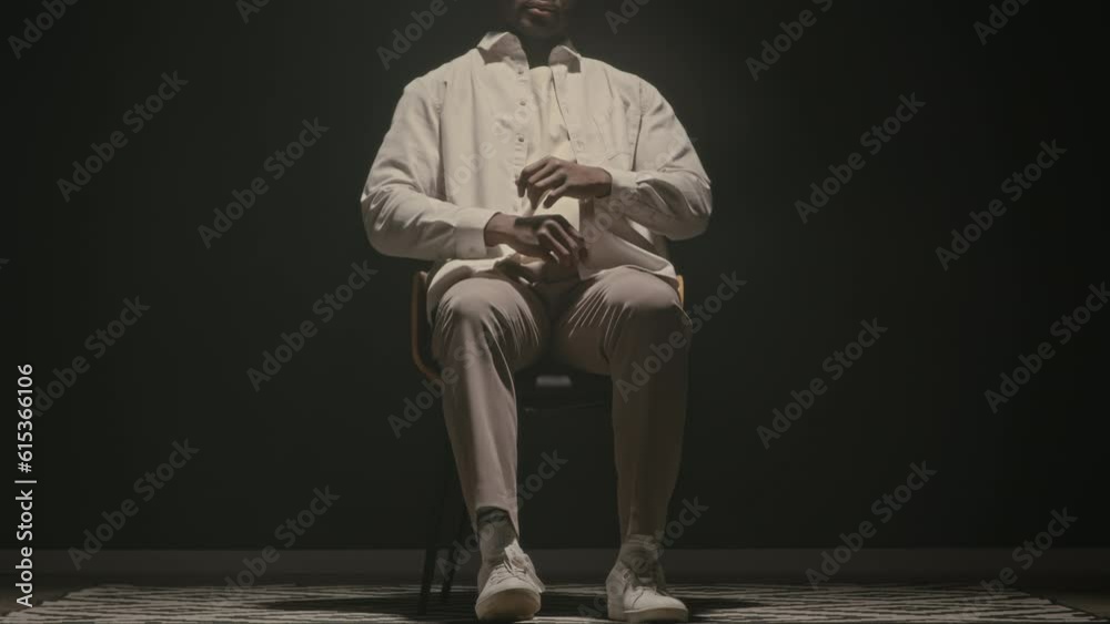 Cropped stab studio portrait of Black man in white clothes sitting down on wooden chair in middle of room on clear black background with dimmed spotlight shining from above
