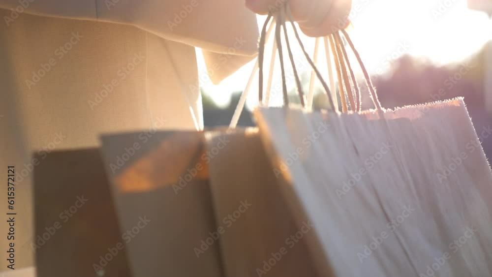 Hands of young woman carry shopping bags walking along urban street. Girl going with paper packets at city square after purchases. Female arm holds packs while going outdoor at sunny day. Close up
