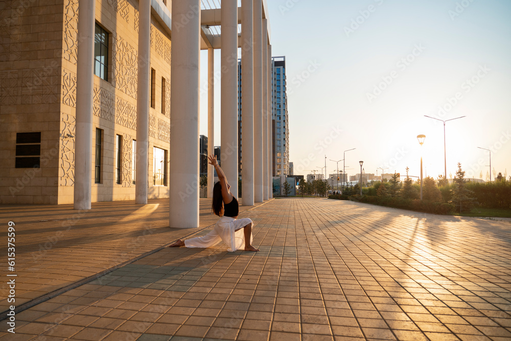 Young Asian woman practicing yoga outdoor during daytime with the view of the city. The girl performs asanas with the view of the columns of the building. Urban yoga, sunrise workout.