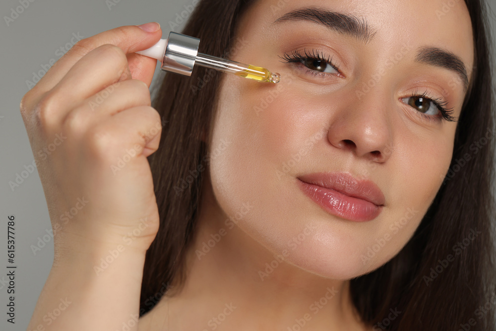 Beautiful young woman applying essential oil onto face on grey background, closeup Beautiful young woman applying essential oil onto face on grey background, closeup