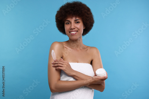 Beautiful young woman applying body cream onto arm on light blue background