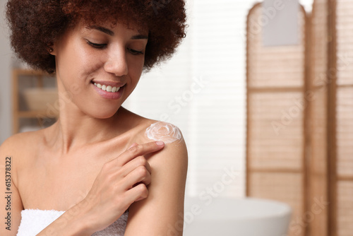 Beautiful young woman applying body cream onto shoulder in bathroom, space for text