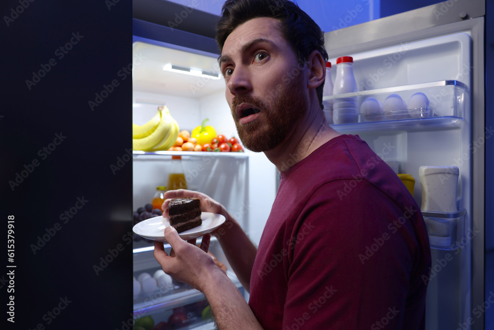 Man taking plate with cake from refrigerator in kitchen at night. Bad habit Man taking plate with cake from refrigerator in kitchen at night. Bad habit
