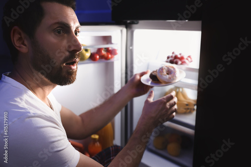 Man taking plate with donuts from refrigerator in kitchen at night. Bad habit