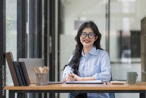 Attractive young Asian businesswoman or female office employee wearing eyeglasses sitting at her office desk, smiling and looking at the camera. © Songsak C
