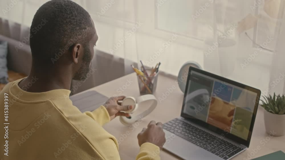 Back view of young adult Black man using laptop computer while working on freelance from home officeBack view of young adult Black man using laptop computer while working on freelance from home office