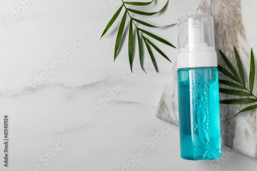 Bottle of face cleansing product and green leaves on white marble table, flat lay. Space for text