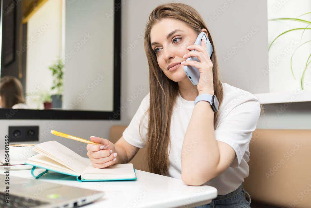 A freelancer girl talks on the phone, writes in a notebook and uses a laptop in a cafe. Freelance concept. A freelancer girl talks on the phone, writes in a notebook and uses a laptop in a cafe. Freelance concept.