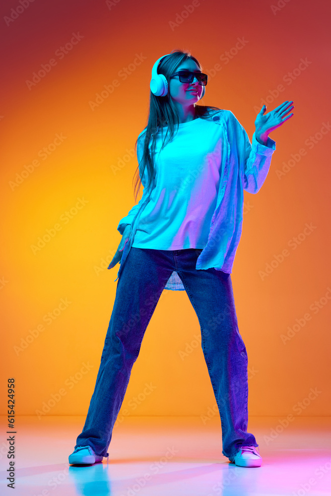 Fototapeta premium Young woman in casual clothes and sunglasses listening to music in headphones and dancing against gradient orange studio background in neon light