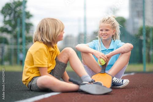  pickleball game, relaxing pickleball players kids boy and girl with yellow ball and paddles sitting after game, outdoor sport leisure activity.