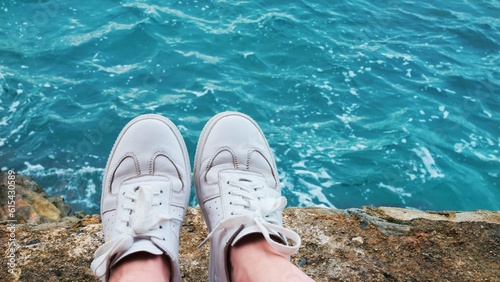 First person view of person sitting to ocean. Point of view young unrecognizable person legs in white shoes cliff sea nature. Traveller in white shoes sitting on rocks, rocky seaside or riverside.