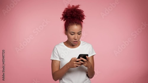 Upset serious curly redhead African American woman using smartphone typing browsing looks camera. Emotional sad female holding mobile cell phone on isolated pink background. People emotions lifestyle