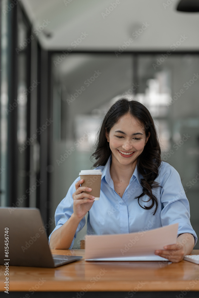 Asian businesswoman holding cup of coffee and financial business papers sitting resting while working using laptop, calculator to analyze investment finance report, startup business concept.