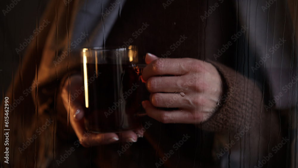 Close-up of glass cup with tea in female hands outside window wet from rain,drops flow down glass.Atmospheric photo autumn mood sadness,melancholy, loneliness.Autumn,fall,home comfort in cold season.