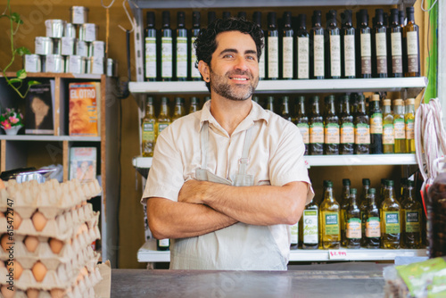 Latin man greengrocer in an organic grocery shop behind the counter with his arms crossed looking at camera