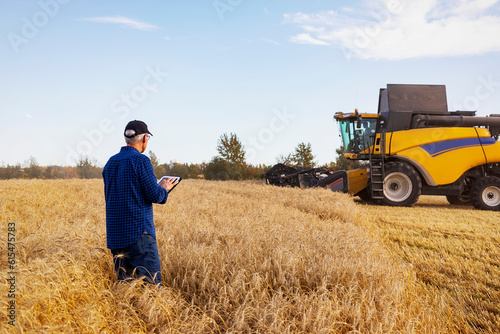 Farmer using a tablet to manage his grain harvest with a combine working in the background; Alcomdale, Alberta, Canada photo