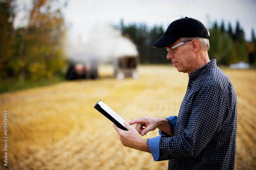 Farmer using a tablet to manage his harvest with harvesting equipment working in the background; Alcomdale, Alberta, Canada Farmer using a tablet to manage his harvest with harvesting equipment working in the background; Alcomdale, Alberta, Canada