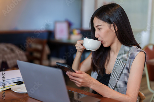 Close-up young Asian woman sitting on mobile phone expression looked cheerful happy, moved hand grab coffee mug that was placed on table and drink break from working on laptop computer, in cafe.