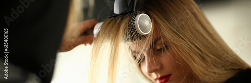 Drying blond hair with a hairdryer and a round brush in barbershop. Master doing hair styling to a client in beauty salon