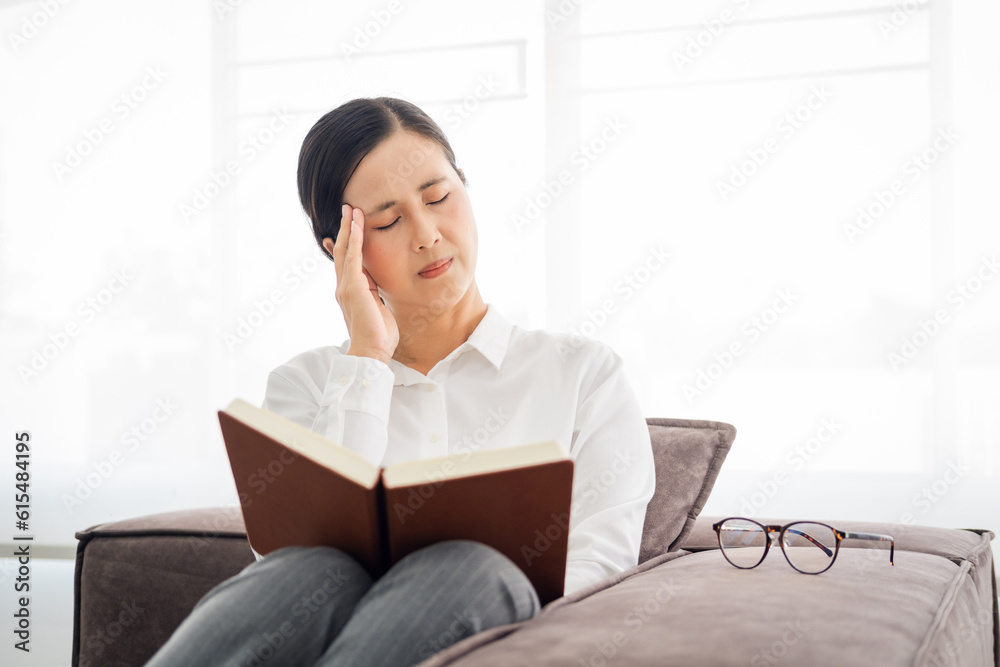 Indoor image of Asian young adult woman in white shirt sitting in living room and having bad headache while reading a book and holding her head out of headache . Indoor image of Asian young adult woman in white shirt sitting in living room and having bad headache while reading a book and holding her head out of headache .