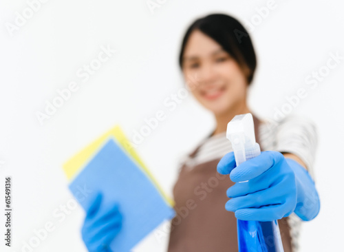 Housekeeper woman holding blue bottle with cleaner liquid and sponge in hands, female wearing white t shirt ,brow apron and blue gloves, looking smiling directly at camera, posing against white wall.