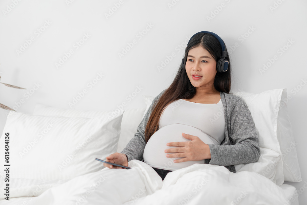 Asian happy pregnant woman is sitting on bed and and touching her belly and wear a headphone to listen the music. pregnancy, motherhood, people and expectation concept Asian happy pregnant woman is sitting on bed and and touching her belly and wear a headphone to listen the music. pregnancy, motherhood, people and expectation concept
