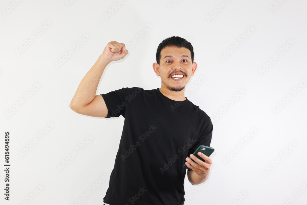 Adult Asian man clenched fist while holding smartphone and showing happy and enthusiastic expression Adult Asian man clenched fist while holding smartphone and showing happy and enthusiastic expression