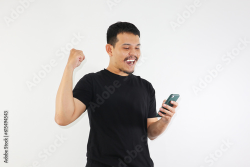 Adult Asian man clenched fist while holding smartphone and showing happy and enthusiastic expression photo