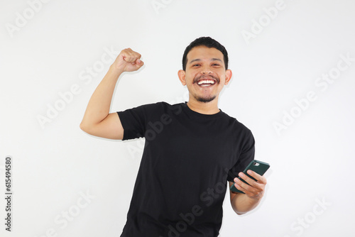 Adult Asian man clenched fist while holding smartphone and showing happy and enthusiastic expression photo